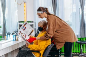 Side view female master in medical mask and gloves showing album with hairstyles images to client on chair in salon